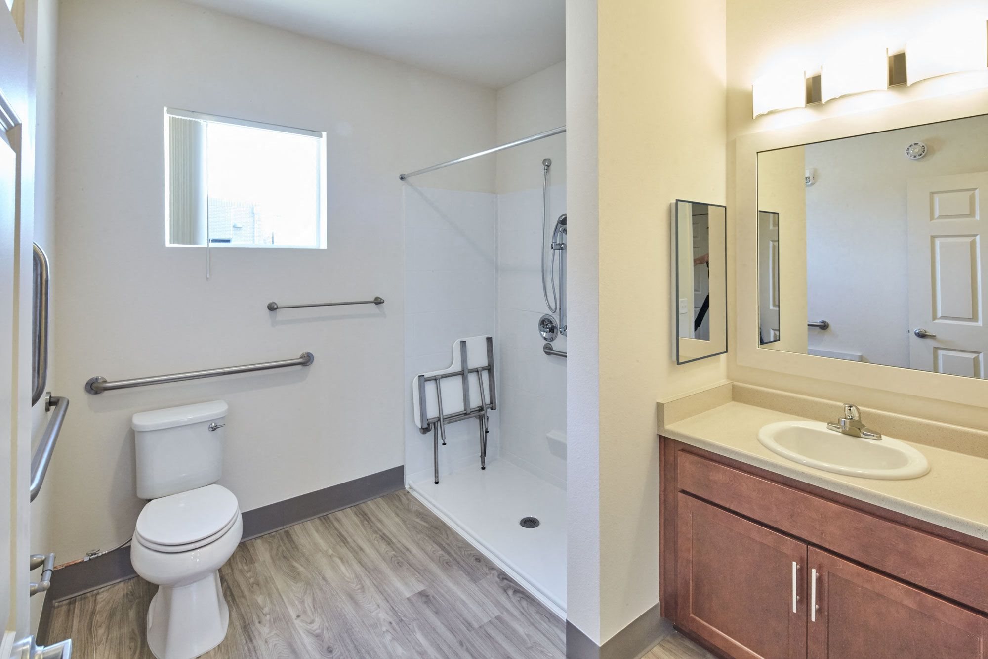 ADA compliant bathroom with walk in shower with attached foldable seat, brown cabinet under sink, and wood like flooring